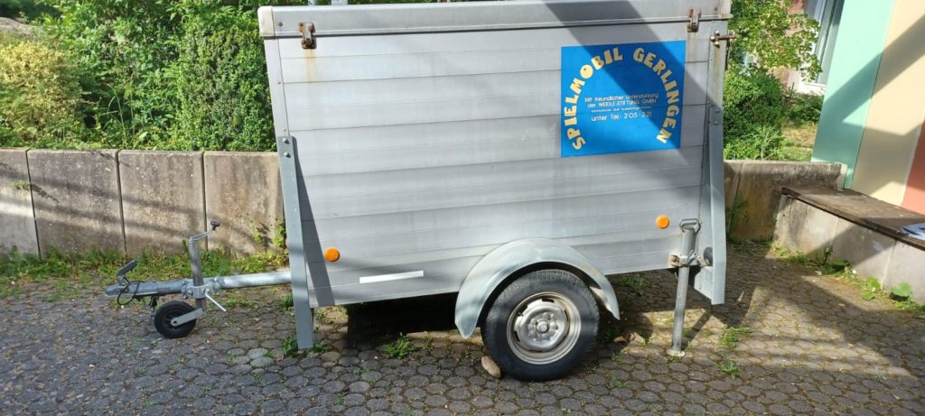 Zu sehen ist ein kleiner Anhänger für Autos mit einem blauen Aufkleber auf welchem in gelber Schrift steht "Spielmobil Gerlingen". Darunter steht kleiner geschrieben: "mit freundlicher Unterstützung der Weidle-Stiftung GmbH"