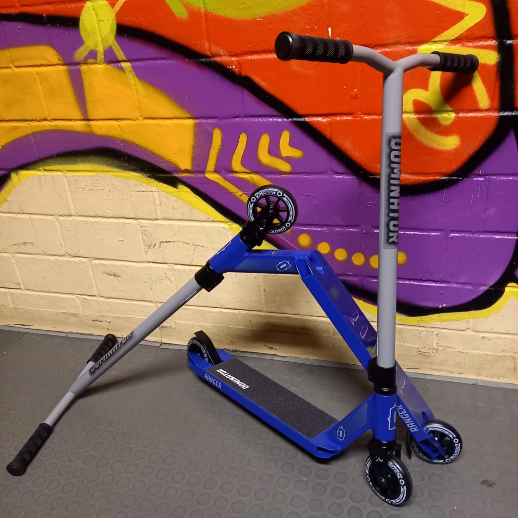 Zu sehen sind zwei Stunt-Scooter von denen einer auf den Rollen steht und der andere umgedreht auf der hinteren Rolle und auf dem Lenker steht. Sie sind vor einer mit Graffiti verzierten Wand im Jugendhaus.